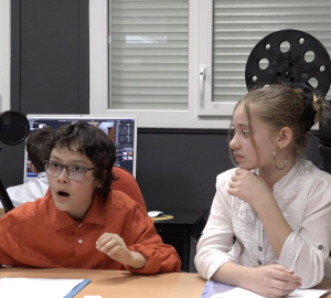 Assis derrière une table et entre deux caméras, un garçon et une fille qui le regarde en faisant la mouee que le ragerde