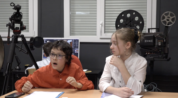 Assis derrière une table et entre deux caméras, un garçon et une fille qui le regarde en faisant la mouee que le ragerde